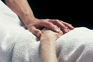 Therapeut einer für Psychosomatik spezialisierten Klinik hält die Hand eines bettlägerigen Patienten. Eine bildliche Einleitung für das Thema der Seite.